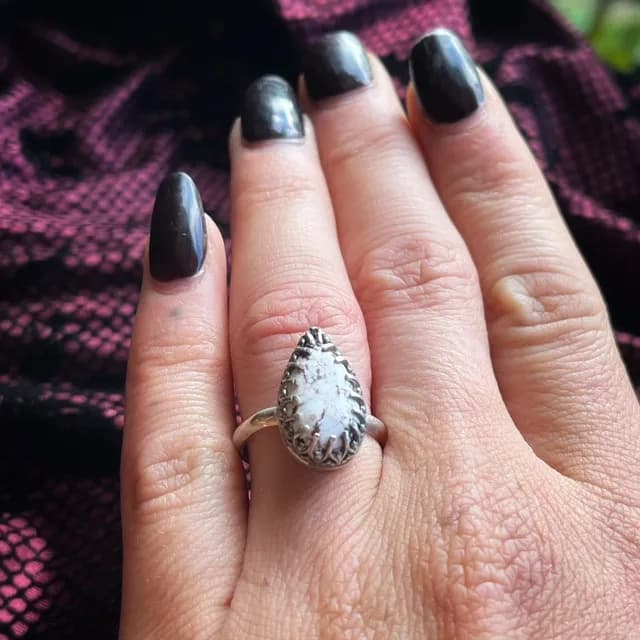 Bague avec pierre en forme de goutte portée à l'annulaire d'une main aux ongles vernis de noir.