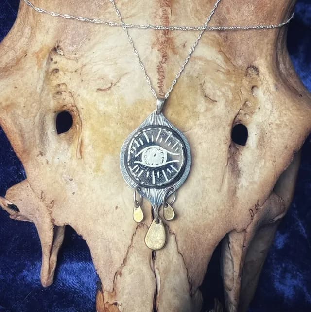 Gros plan sur le pendentif Œil de sorcière gravé à pampilles dorées sur fond texturé.