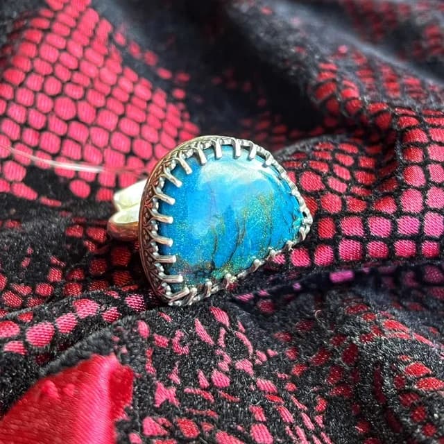 Gros plan sur la bague sertie de sa pierre chrysocolle bleue aux reflets nuancés sur fond rose et noir.