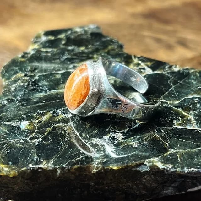 Chevalière en argent avec pierre de soleil orangée et motif gravé sur le côté, posée sur une roche sombre.
