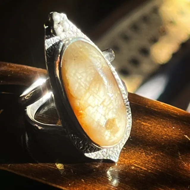 Bague argentée ornée d'un grand cabochon ovale beige doré sur support en bois au soleil