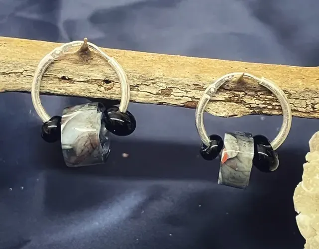Paire de créoles argentées avec perles en verre marbré et perles noires présentées sur une branche de bois.