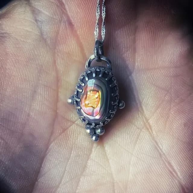 Pendentif en argent patiné serti d'un cabochon d'abalone aux reflets dorés et orangés, tenu au creux d'une main.
