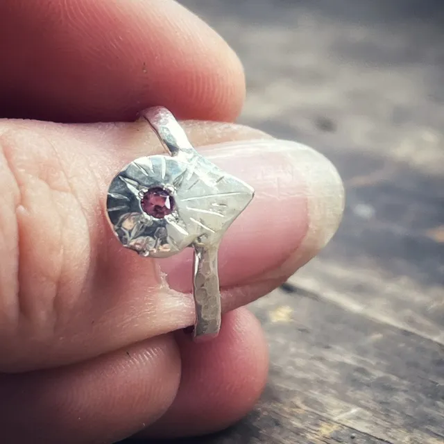 Bague en argent gravé tenue entre les doigts, mettant en valeur une petite pierre rose centrale.