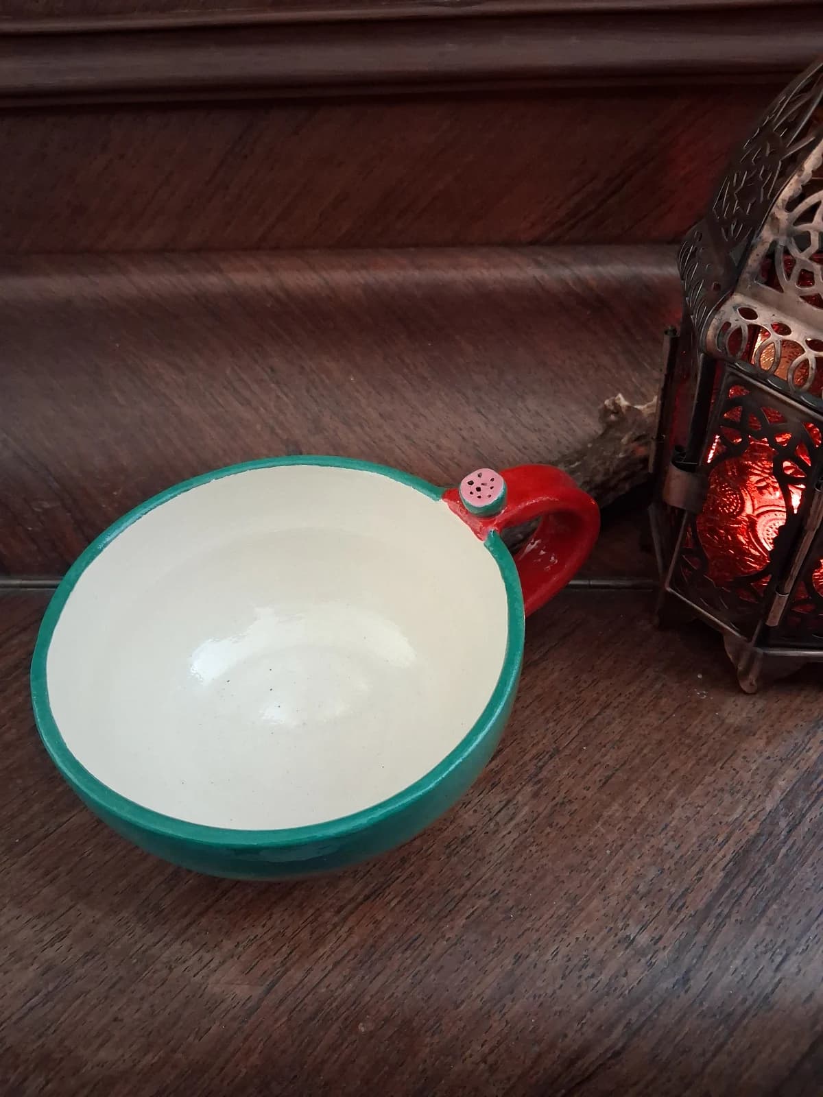 Tasse en céramique verte et crème avec anse rouge ornée d'un détail pastèque, posée sur un meuble en bois.
