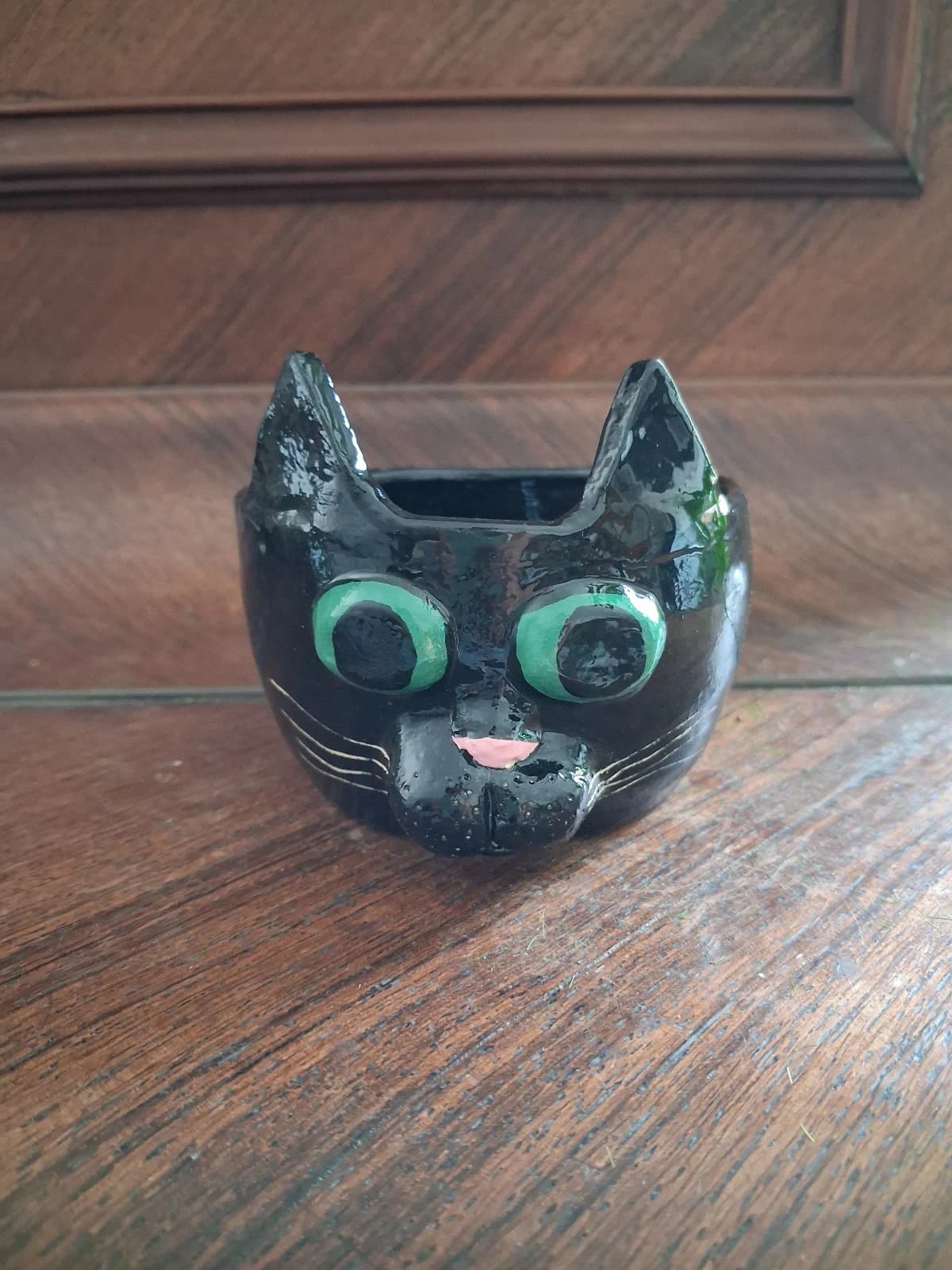 Pot en céramique noire en forme de tête de chat aux yeux verts et nez rose, posé sur un meuble en bois.