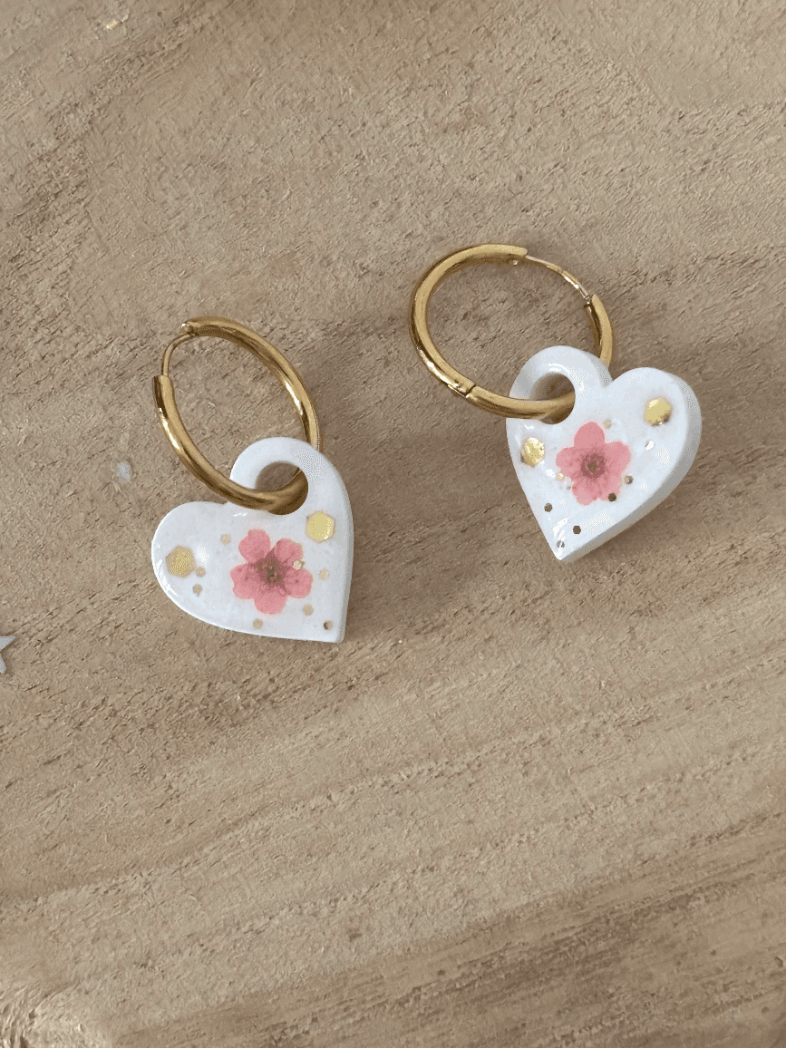 Anneaux dorés avec pendentifs cœurs blancs ornés d'une fleur rose et de paillettes dorées posés sur du bois.