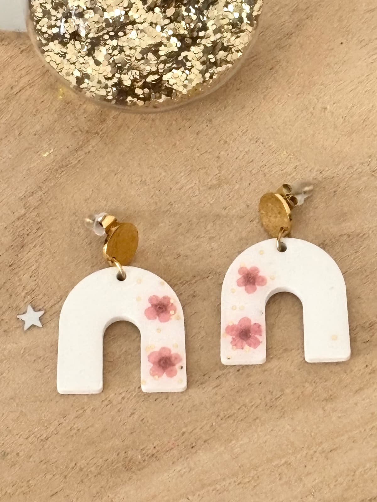 Boucles d'oreilles blanches en forme d'arche avec fleurs roses et puces dorées posées sur du bois.