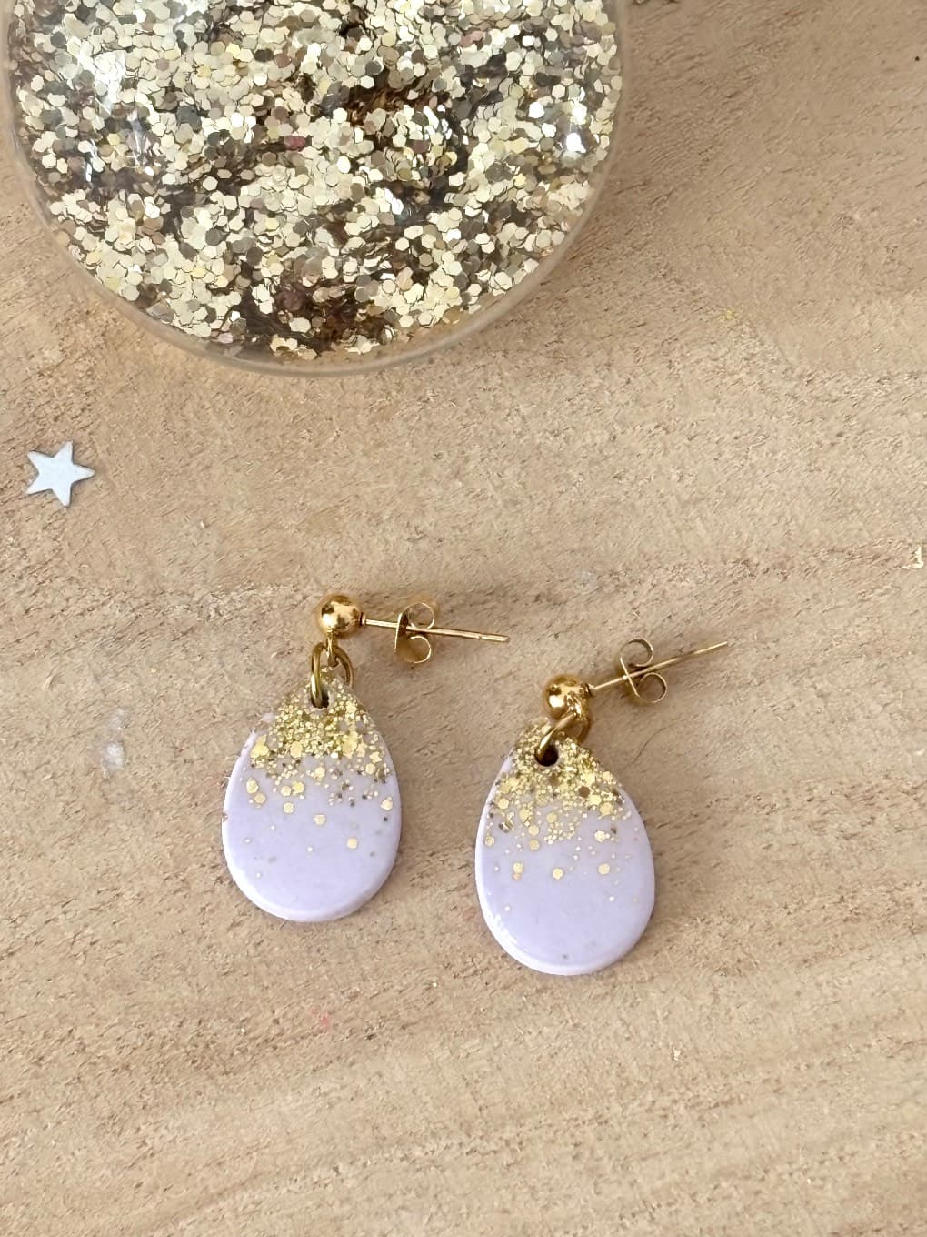 Paire de boucles d'oreilles lilas et dorées posée sur bois près d'un récipient de paillettes