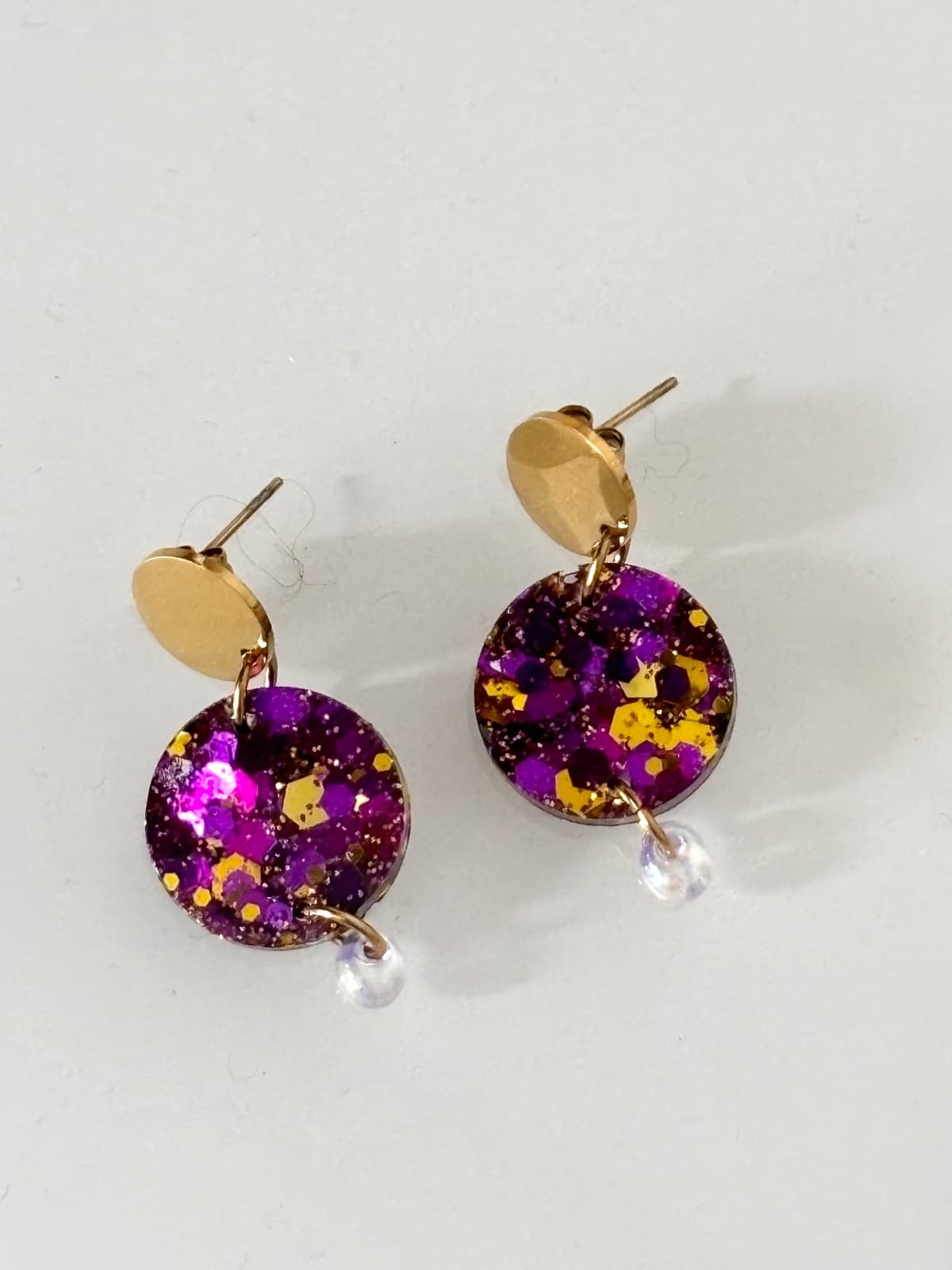 Paire de boucles d'oreilles pendantes rondes violettes et dorées pailletées sur fond blanc.