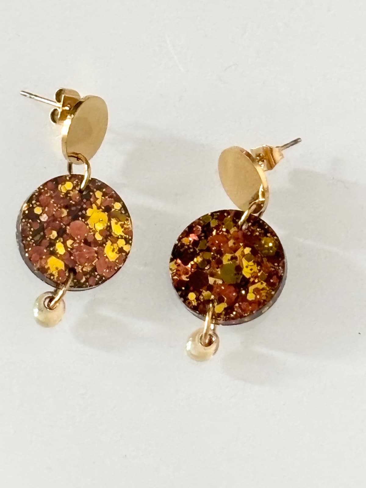 Boucles d'oreilles dorées avec médaillon marron scintillant et perle ambre sur fond blanc.