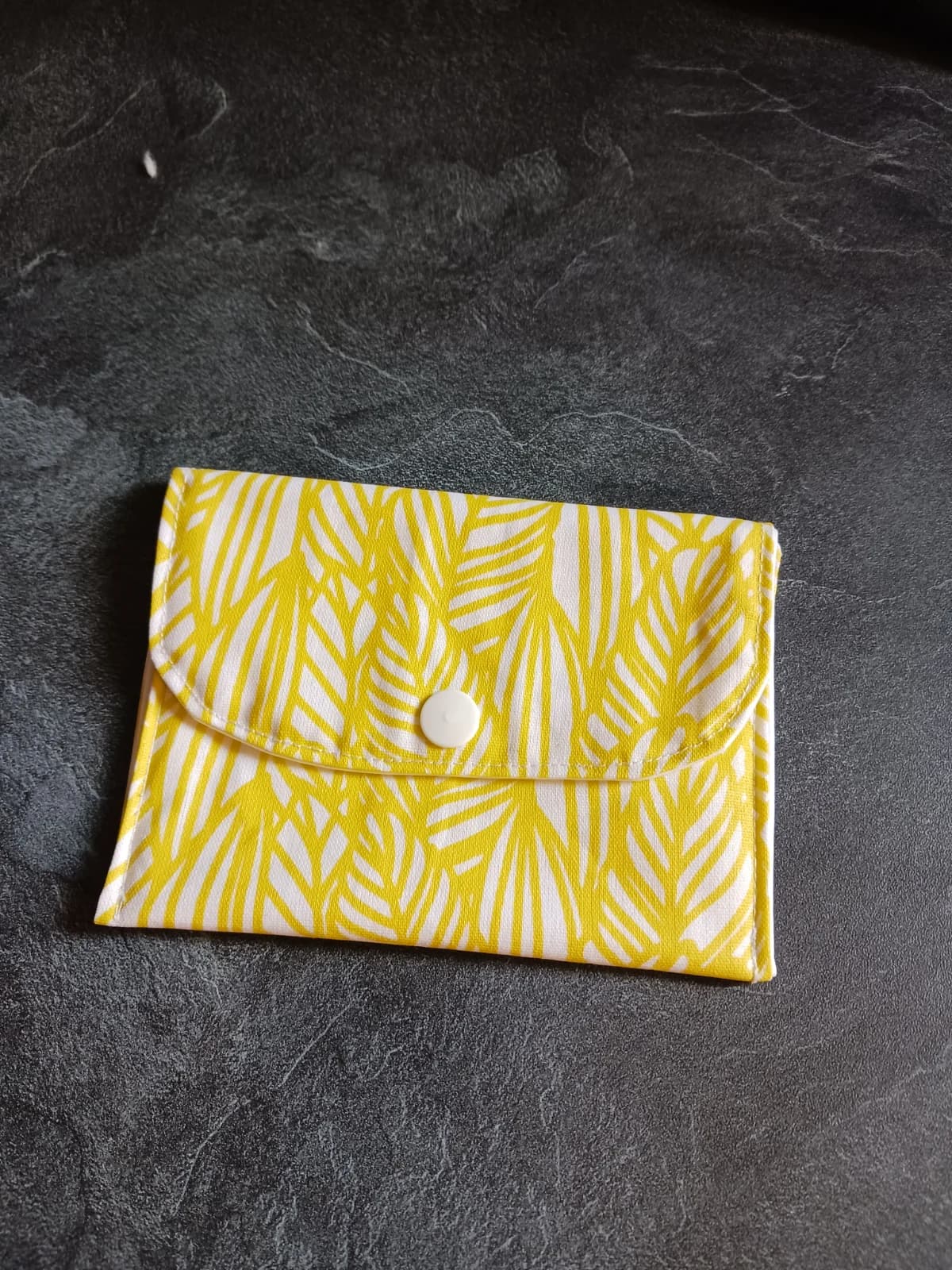 Pochette fermée en tissu à imprimé végétal jaune et blanc avec rabat et bouton-pression blanc.