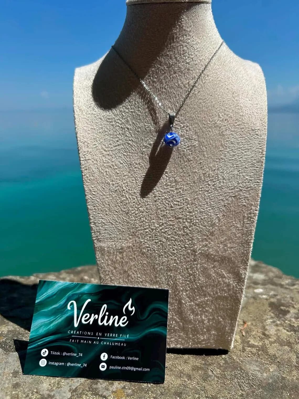 Gros plan sur le pendentif perle en verre filé bleu et blanc sur une fine chaîne argentée.