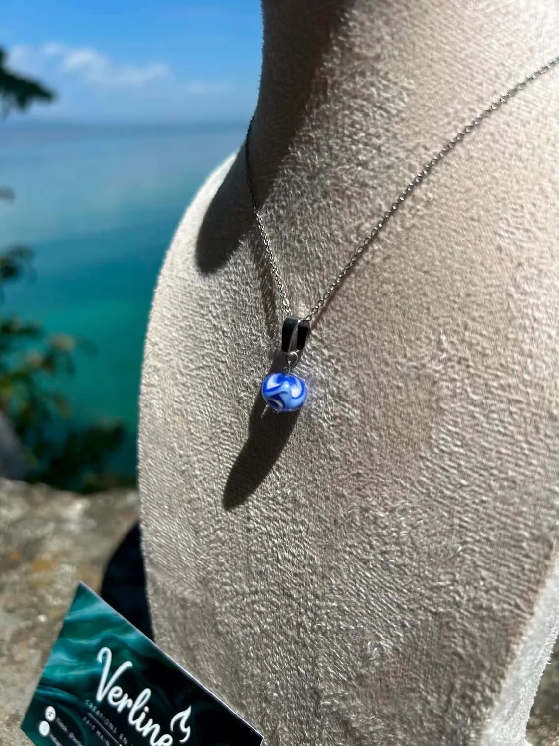 Collier avec pendentif perle ronde bleue et blanche présenté sur un buste face à la mer.
