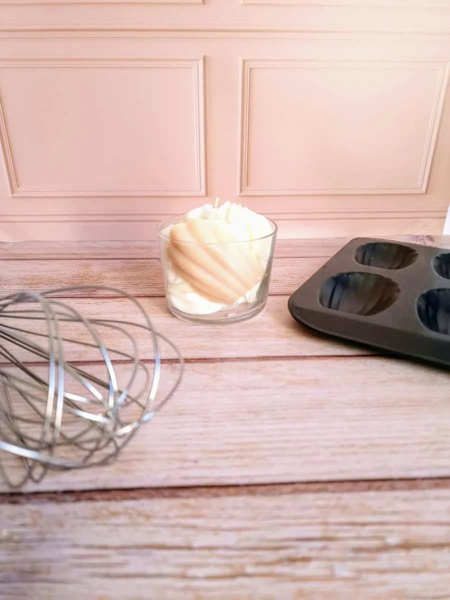Bougie dans un verre transparent décorée d'une madeleine en cire beige, posée près d'un fouet et d'un moule.