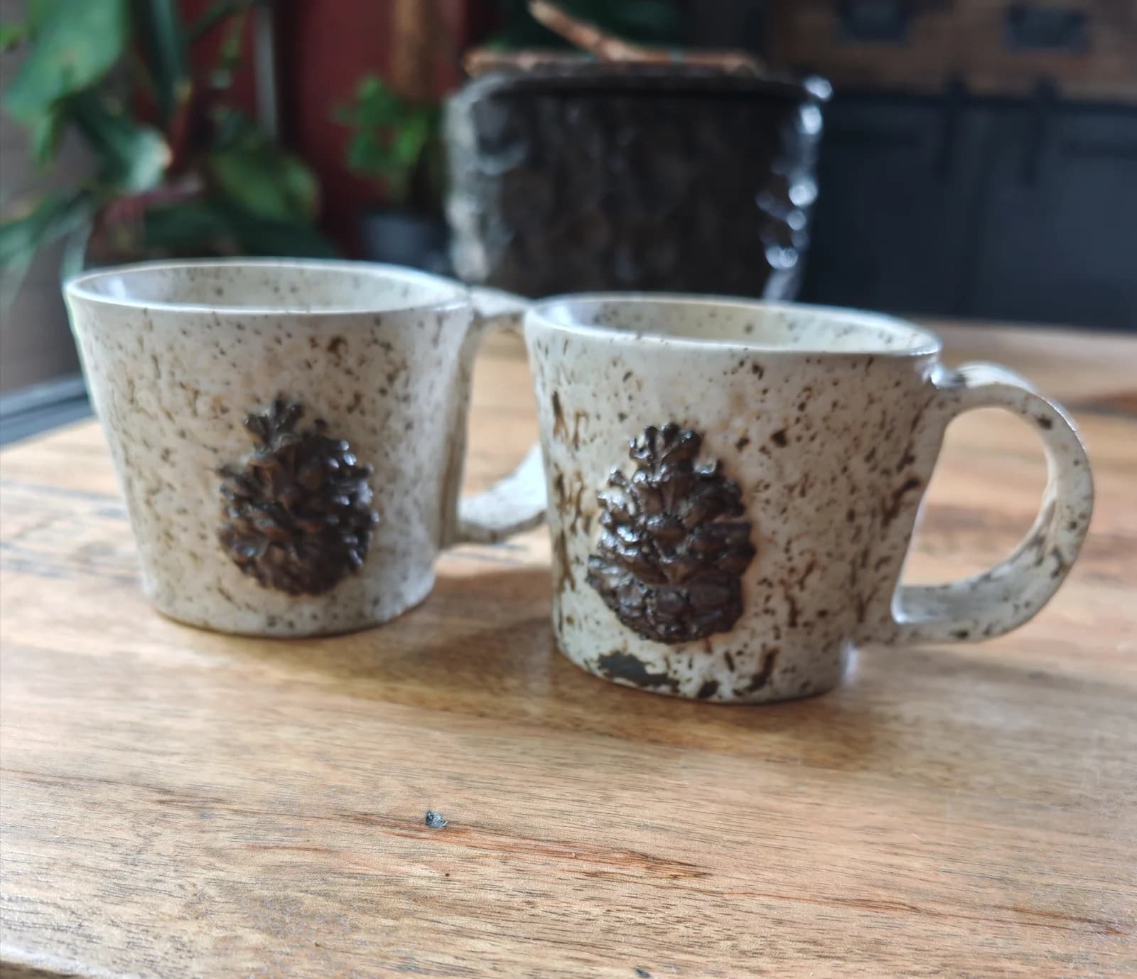 Deux tasses en céramique mouchetée avec motif pomme de pin en relief, posées sur une table en bois.