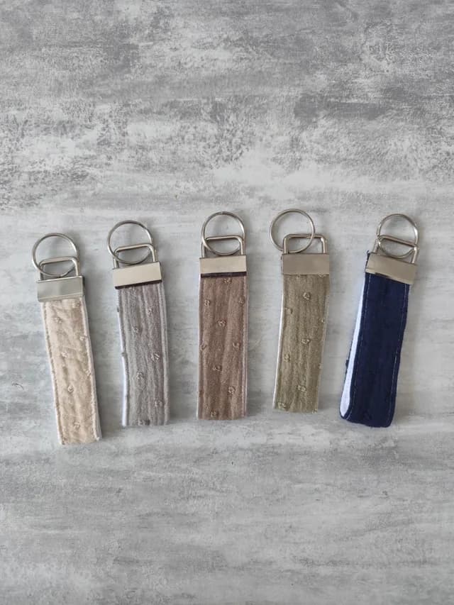 Alignement de cinq porte-clés en tissu texturé beige, gris, marron, kaki et bleu avec boucle en métal