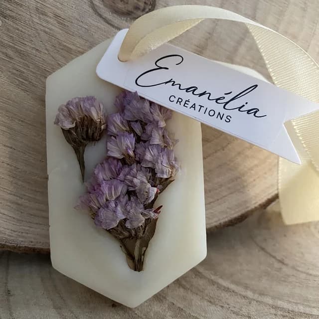 Suspension hexagonale en cire blanche ornée de fleurs séchées violettes, munie d'un ruban clair.