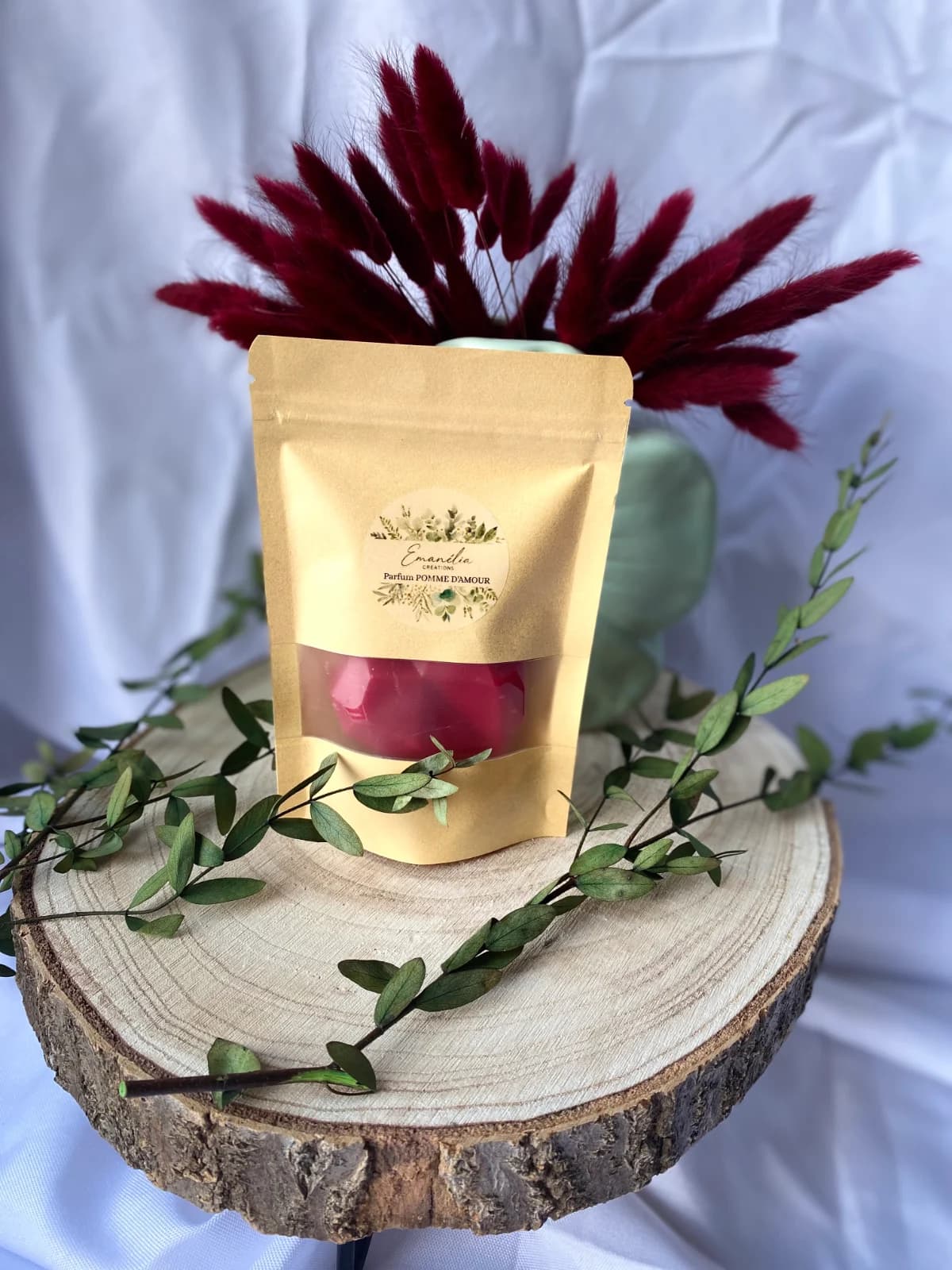 Sachet kraft avec fenêtre laissant voir un fondant rouge, posé sur un rondin de bois décoré de feuillage.