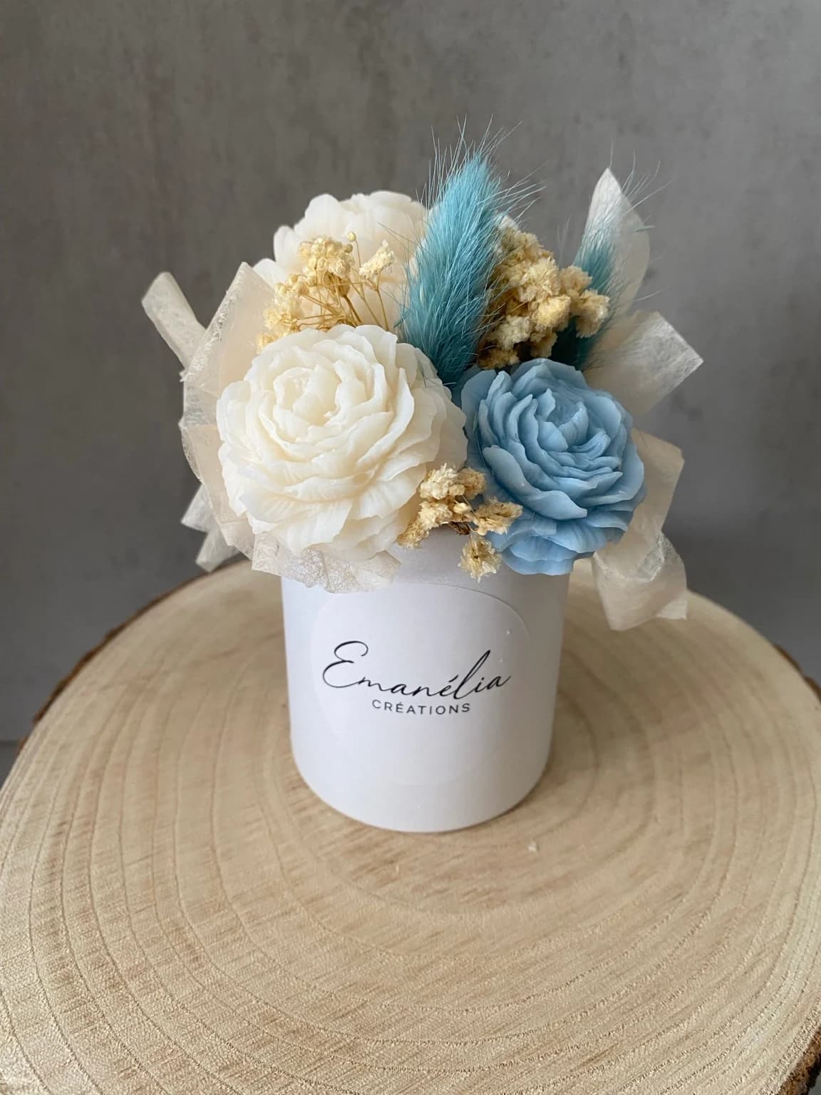 Bouquet de fleurs sculptées blanc ivoire et bleu poudré dans un petit pot blanc posé sur un rondin.
