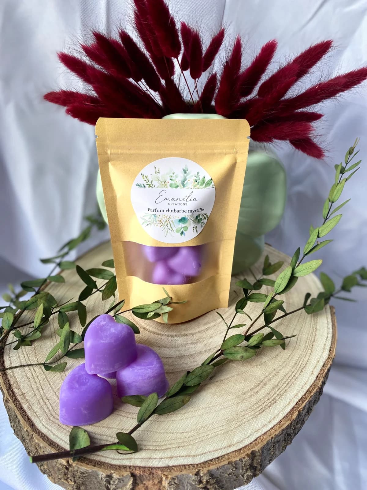Sachet kraft et fondants violets en forme de cœur posés sur une tranche de bois ornée de feuillage.